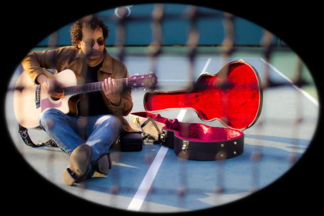 a man sitting on tennis court playing guitar alongside is his guitar case